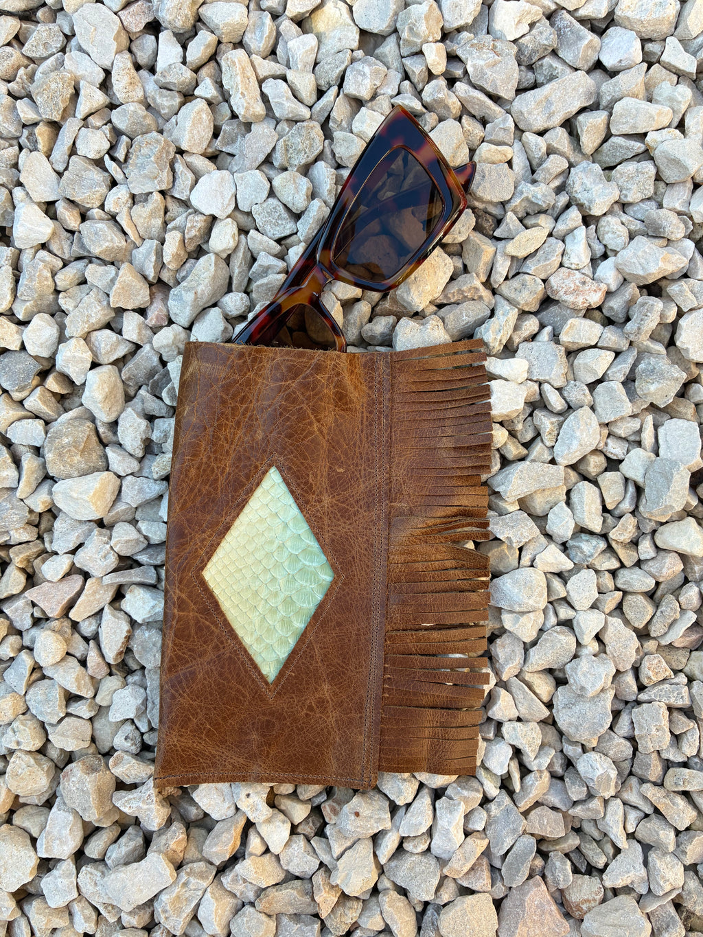 A brown leather sunglasses holder with fringe and a diamond-shaped textured inlay, shown on a bed of small gray stones with tortoiseshell sunglasses resting inside.