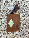 A brown leather sunglasses holder with fringe and a diamond-shaped textured inlay, shown on a bed of small gray stones with tortoiseshell sunglasses resting inside.