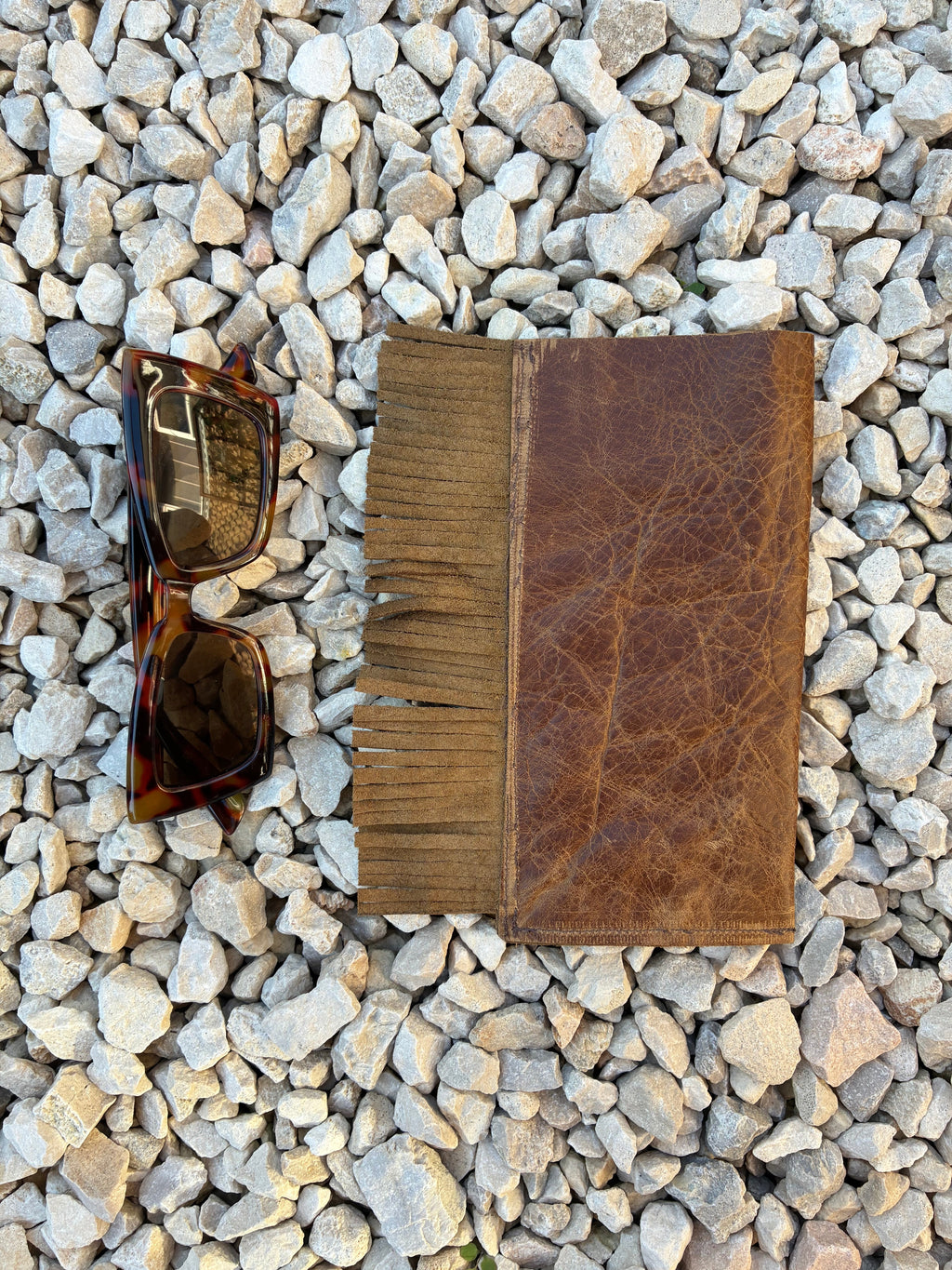 A brown leather glasses case with fringe detailing across the top, displayed on a bed of small gray pebbles next to a pair of tortoiseshell sunglasses.