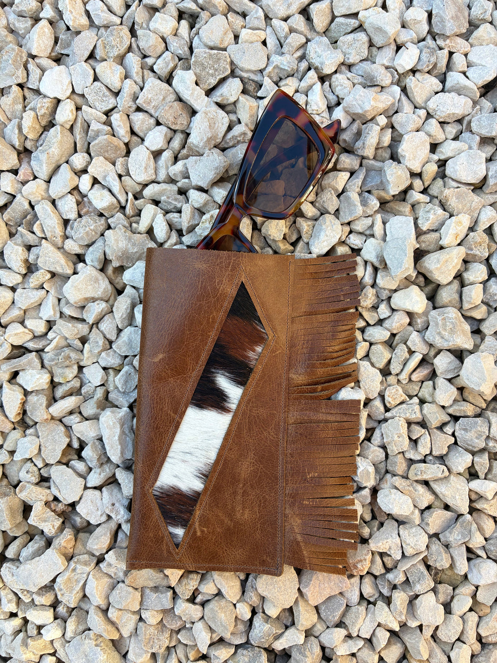 A brown leather glasses case with fringe and a geometric cutout showing a black-and-white cowhide pattern, styled on a bed of light gray pebbles next to tortoiseshell sunglasses.