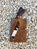 A brown leather glasses case with fringe and a geometric cutout showing a black-and-white cowhide pattern, styled on a bed of light gray pebbles next to tortoiseshell sunglasses.