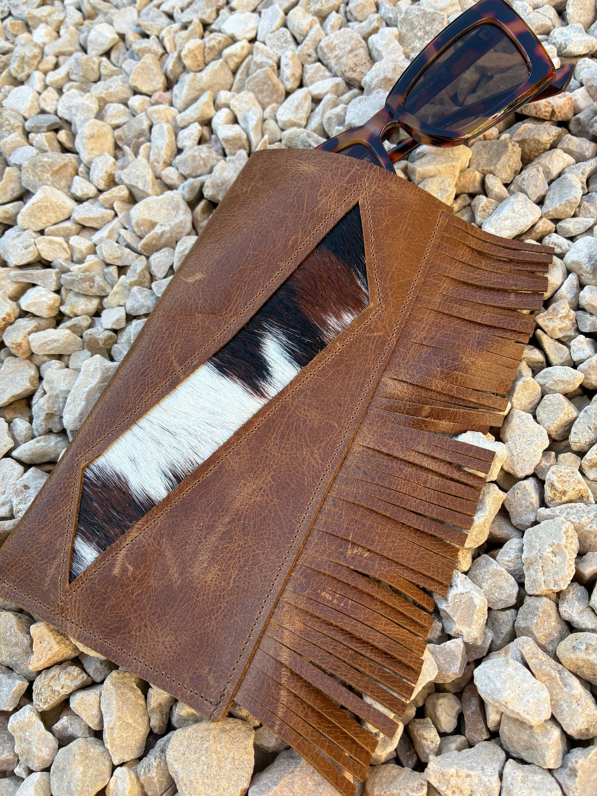 A brown leather glasses case with fringe detailing and a central cowhide patch, styled on a bed of light gray pebbles with sunglasses resting on the corner.