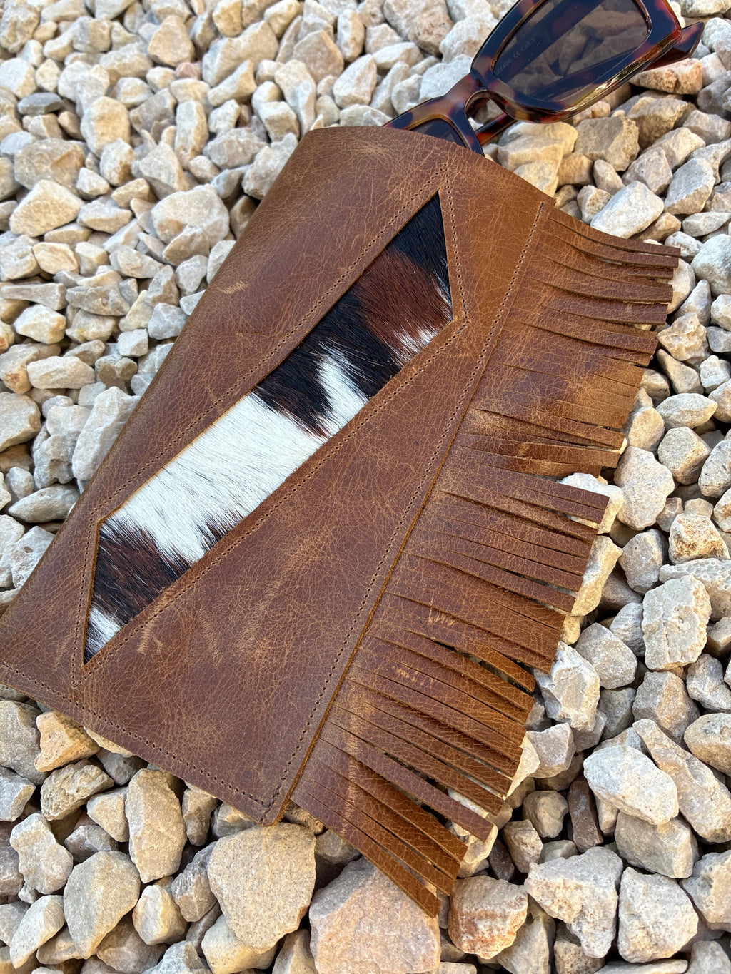 A brown leather fringe glasses case with a cowhide hair panel, laid flat on a bed of small gray stones with sunglasses resting on the corner.