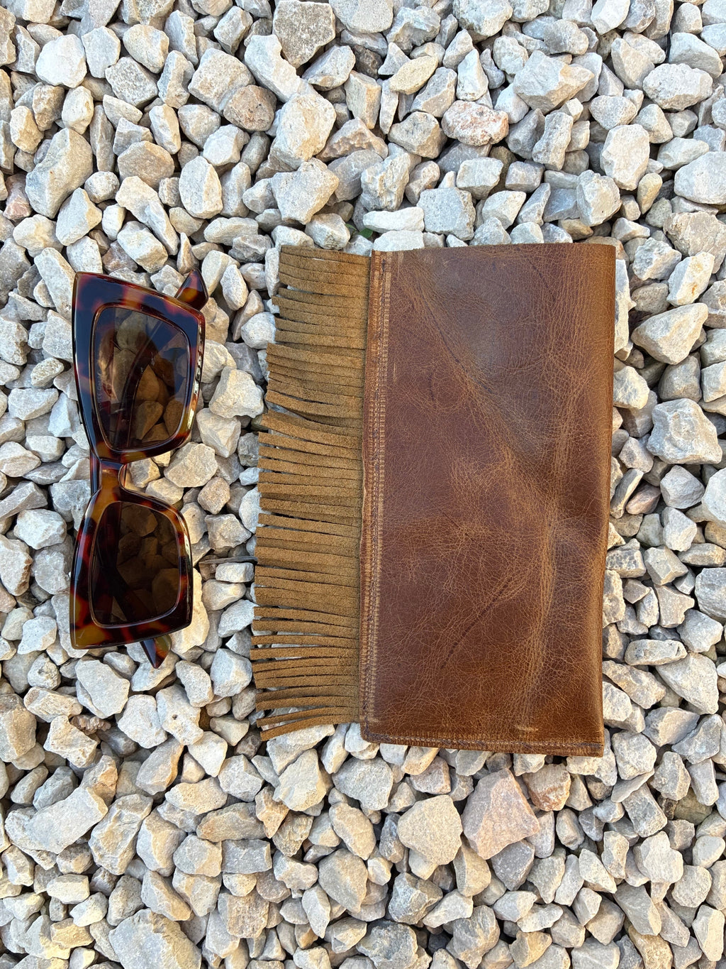 A brown leather glasses case with fringe detailing is laid flat on a bed of small gray pebbles, styled with a pair of tortoiseshell sunglasses above it.