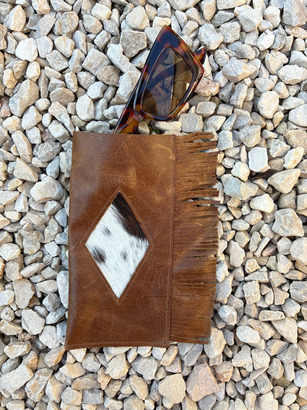 A brown leather glasses case with fringe and a diamond-shaped cowhide patch, styled on a bed of gray pebbles next to tortoiseshell sunglasses.