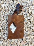 A brown leather glasses case with fringe and a diamond-shaped cowhide patch, styled on a bed of gray pebbles next to tortoiseshell sunglasses.